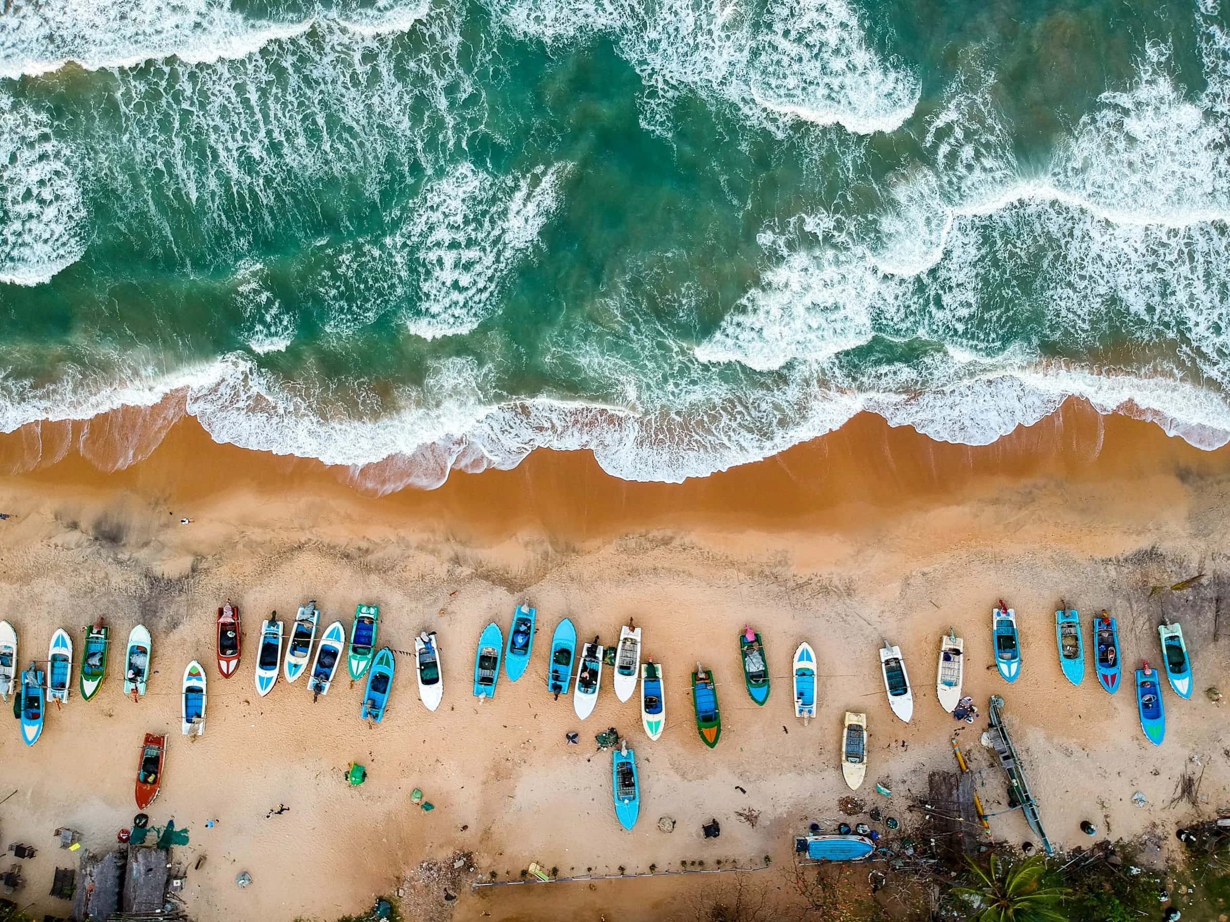Aerial view of fishing boats lined up on a beach beside turquoise surf